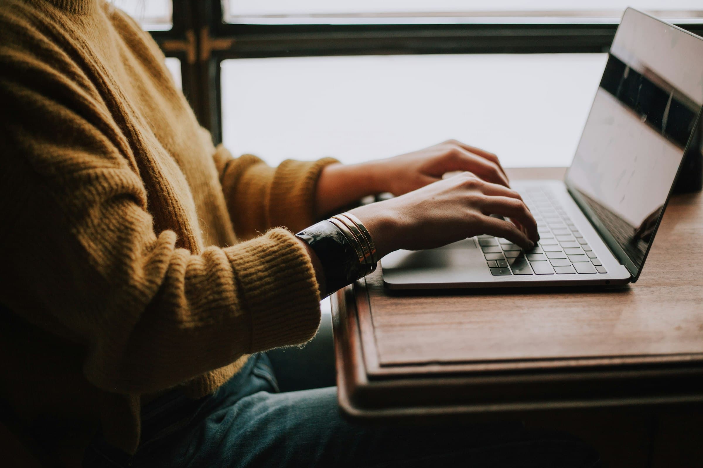 Manos escribiendo en el teclado de una laptop sobre una mesa de madera, junto a una ventana